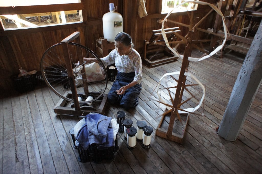 A woman kneels beside a traditional spinning wheel, winding freshly extracted lotus fibre into thread.