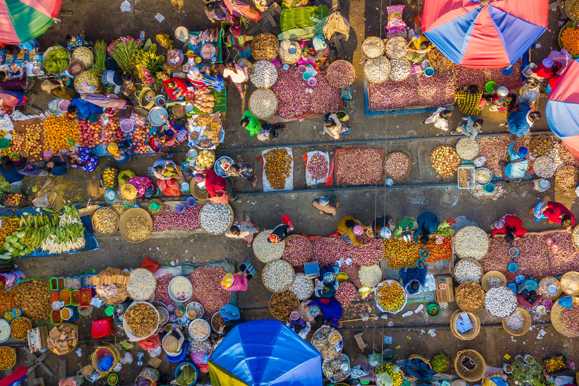 An overhead view of a bustling Myanmar market, with vendors and shoppers arranged along rows of colourful produce, umbrellas and baskets.