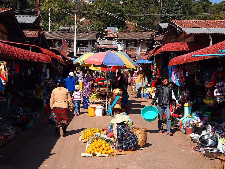 A busy market street lined with red-roofed stalls and striped umbrellas in a Shan State hill town.