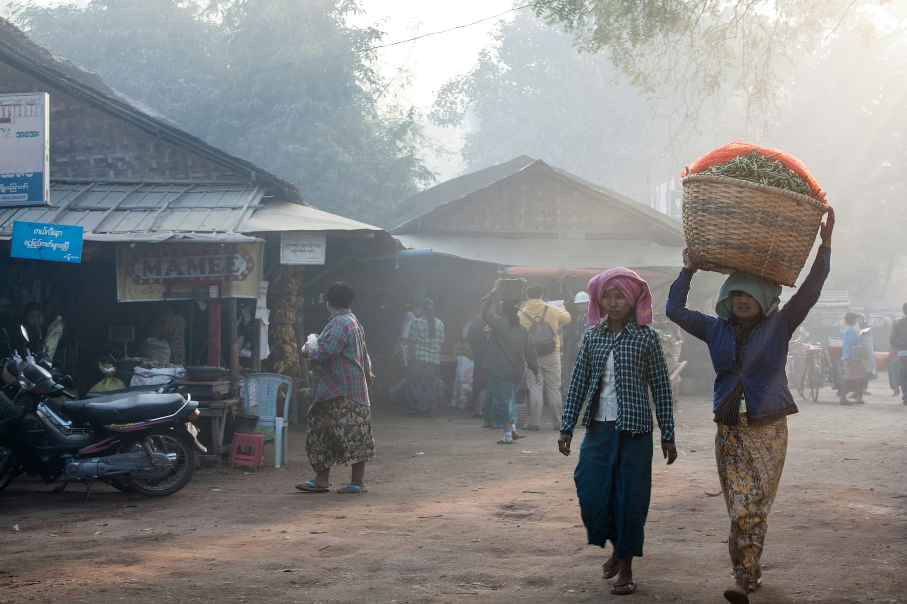 Women carry baskets of produce through a hazy, smoke-filled market street lined with wooden stalls.
