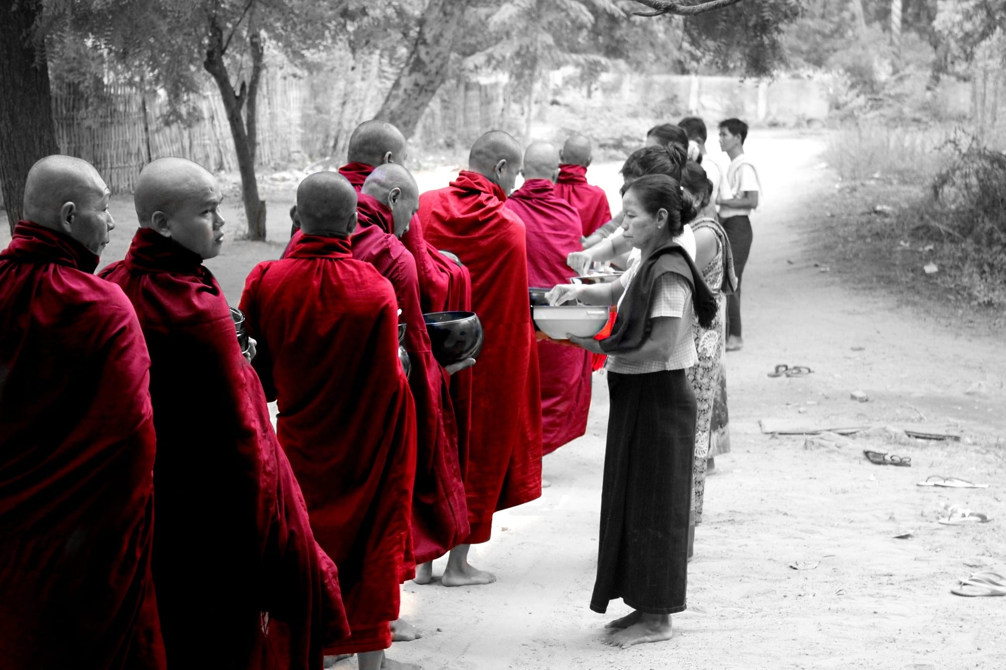 A line of monks in maroon robes, shown in selective colour, receiving alms from women along a village road at dawn.