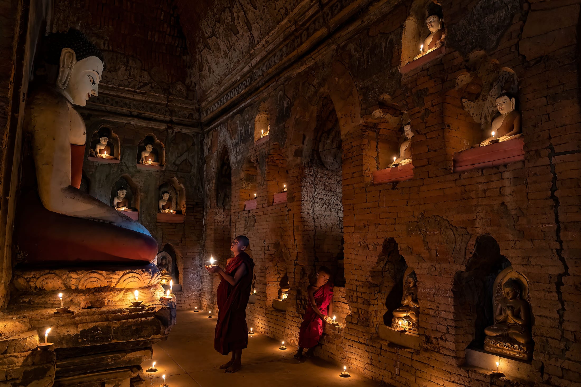Novice monks lighting candles among candlelit Buddha niches inside an ancient Bagan temple