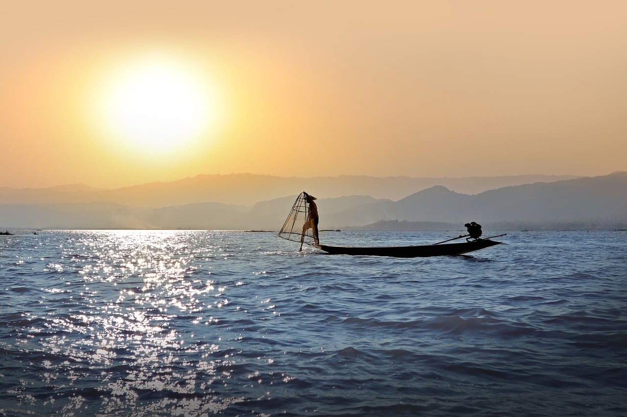 A traditional Inle Lake fisherman balances on one leg to row his boat, silhouetted against a golden sunset over the water