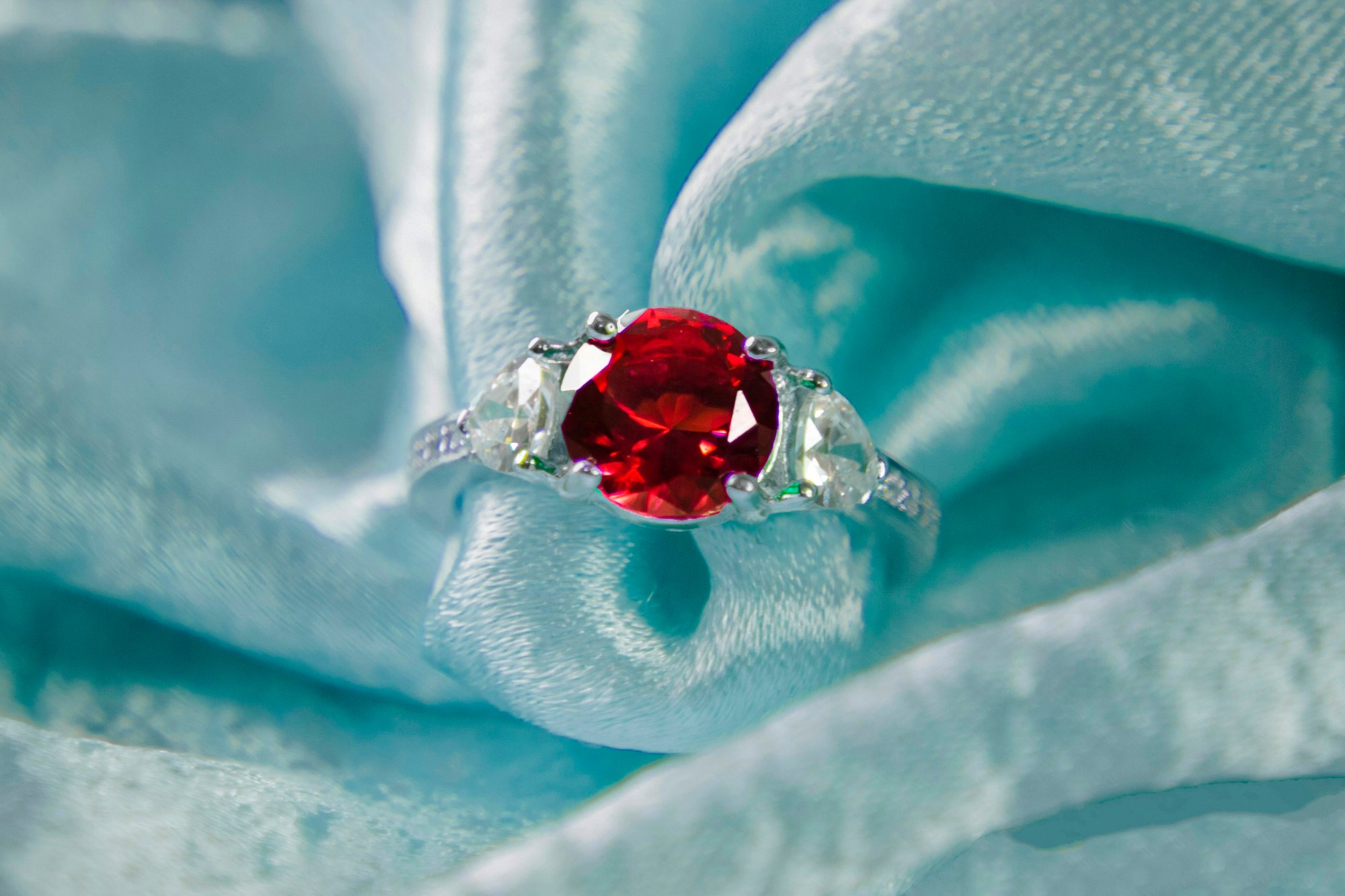 A macro shot of a ruby and diamond ring nestled in folds of teal silk fabric, the deep red stone in sharp focus