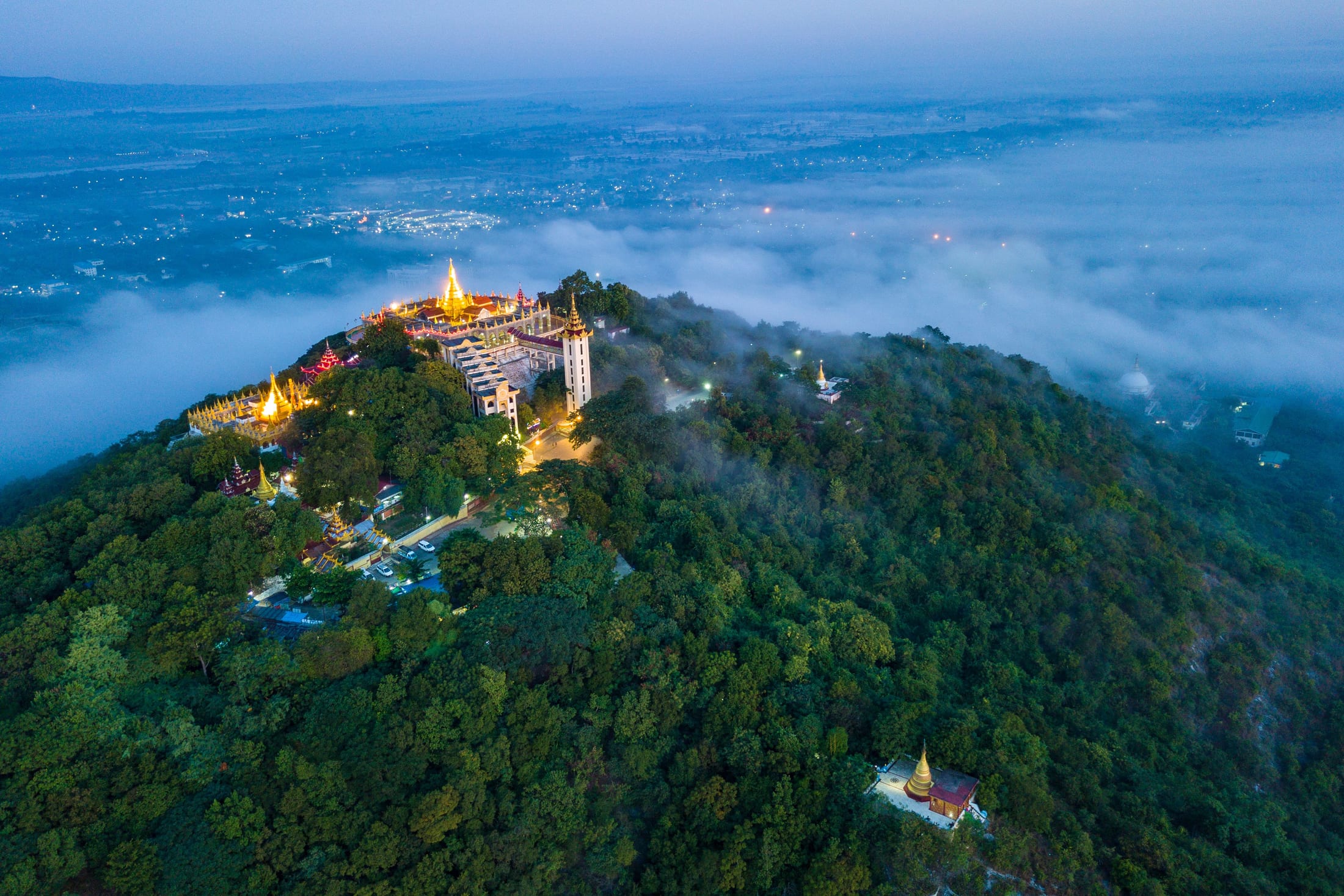 Aerial twilight view of Mandalay Hill's golden pagoda rising above a sea of clouds